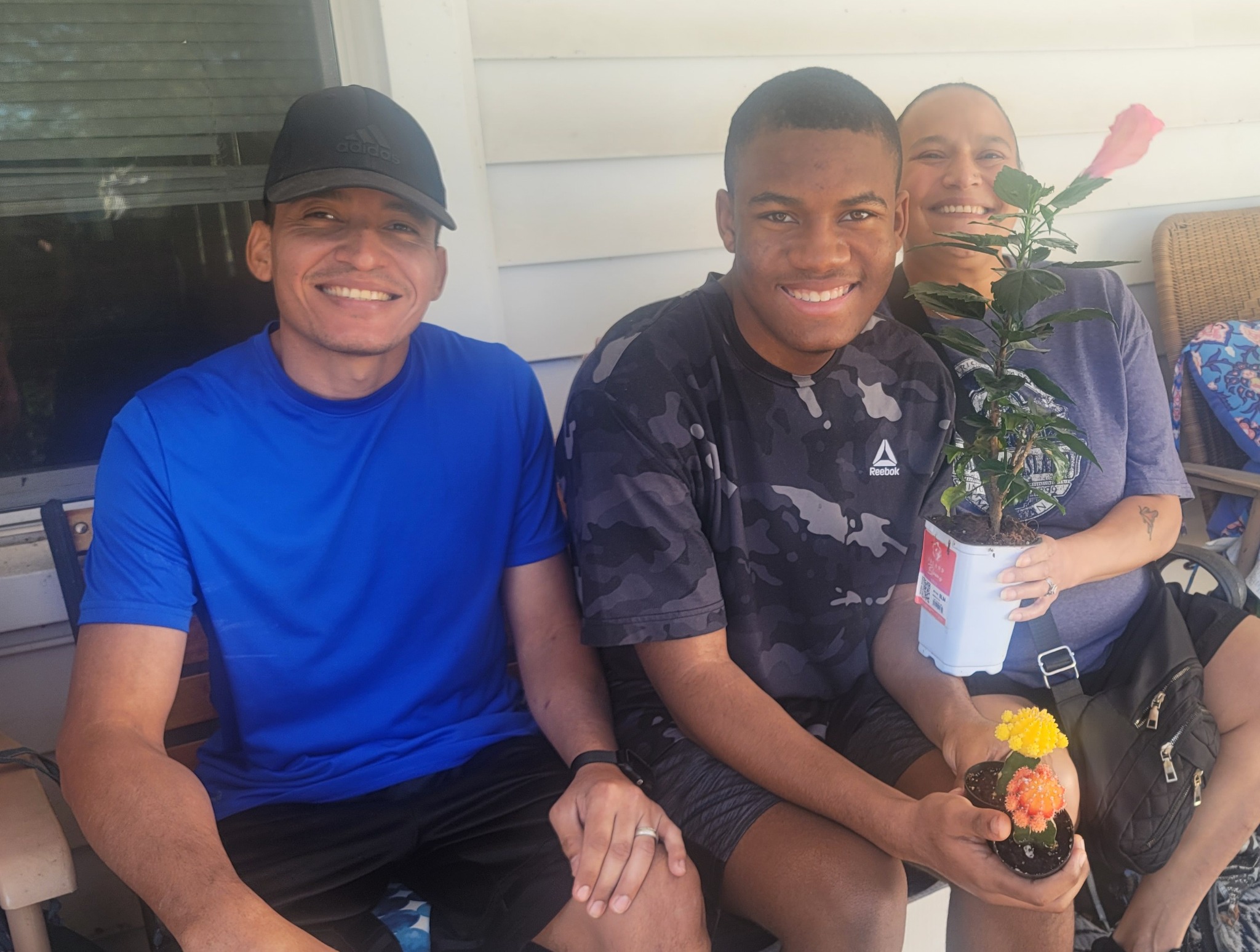 Jelani smiling on a porch with loved ones, holding a flowering cactus beside a pink hibiscus